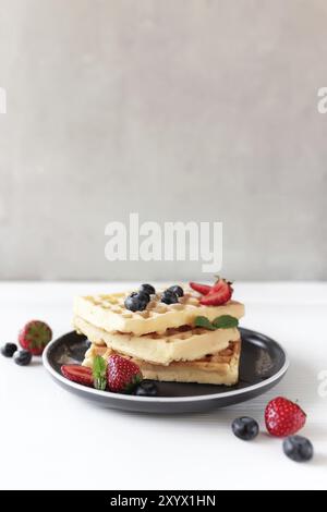 Una pila di waffle su un piatto sul tavolo bianco con mirtilli, fragole tritate e foglie di menta, sfondo grigio. Foto Stock