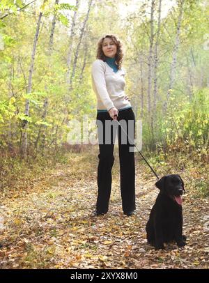 Giovane donna che cammina con il cucciolo nero labrador retriever nella foresta autunnale Foto Stock