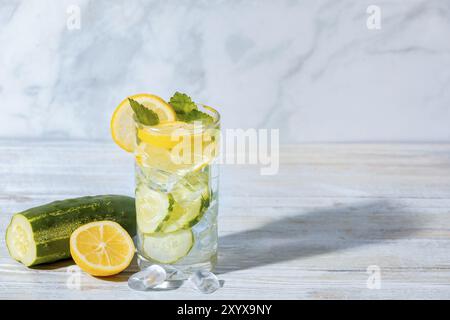 Un bicchiere di acqua cetriolo con fette di limone, foglie di menta e cubetti di ghiaccio, cetriolo e limone su un tavolo accanto ad esso Foto Stock