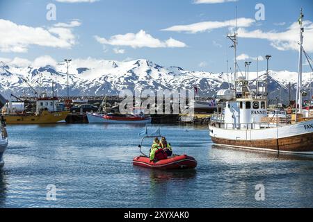 HUSAVIK, ISLANDA, GIUGNO 29: Turisti che tornano da un tour di avvistamento delle balene su uno zodiaco nel tranquillo porto con barca da pesca ancorata e montagne i. Foto Stock