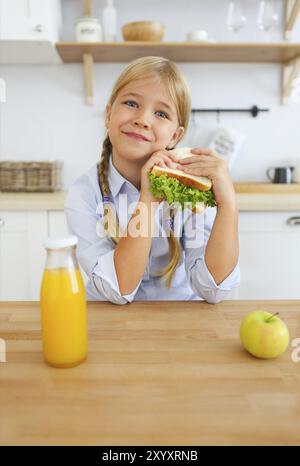 Felice bambina in età scolare, biondo kid godendo di una sana colazione mangiare panino e frutta e bere il succo d'arancia seduti al luminoso ki Foto Stock