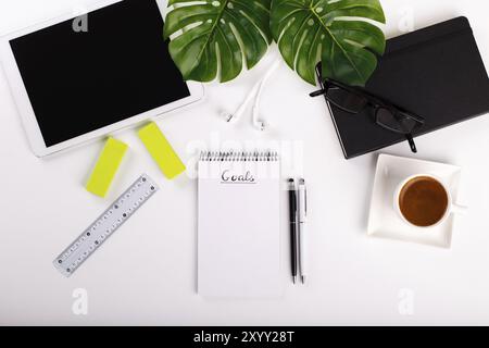Tavolo da ufficio bianco moderno con tablet, occhiali, penna, matita, righello, pianta monstera, blocco note e tazza greca di caffè. Vista dall'alto, lastre piatte cimasa Foto Stock