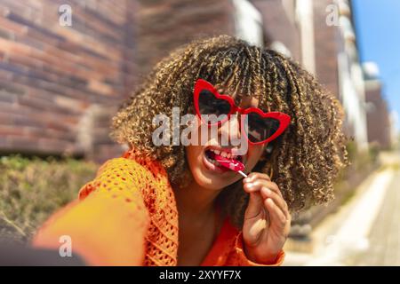 Giovane donna latina che indossa occhiali da sole a forma di cuore, che scatta selfie e mangia lecca lecca Foto Stock