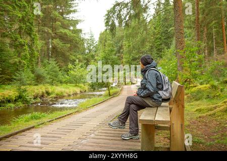 Escursionista/rambler isolata in un abbigliamento impermeabile e all'aperto, seduto su una panchina sul lungomare lungo il fiume nella foresta di Hafren, vicino a Llanidloes, Galles. Foto Stock
