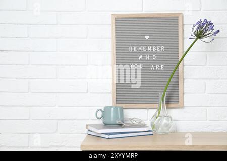 Bacheca con frase che ricorda chi sei, libri, tazze e fiori in vaso su un tavolo in legno all'interno, spazio per il testo Foto Stock