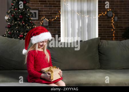 Piccola ragazza sorridente in rosso vestito festivo che tiene regalo di Natale o di Capodanno. Bambino caucasico vestito in cappello o cappello di Santa seduta sopra Foto Stock