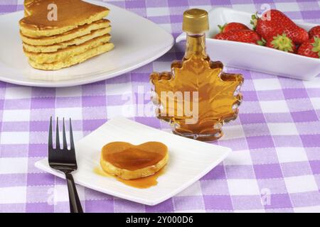 Mini pancake a forma di cuore con sciroppo su un piatto bianco con forchetta. Sullo sfondo una pila di pancake una bottiglia di sciroppo d'acero e alcune fragole. A. Foto Stock