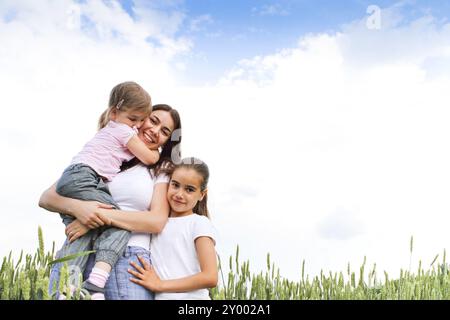 Piacere giovane madre ridere e guardare la macchina fotografica e abbracciando piccole figlie con campo verde e cielo sullo sfondo Foto Stock