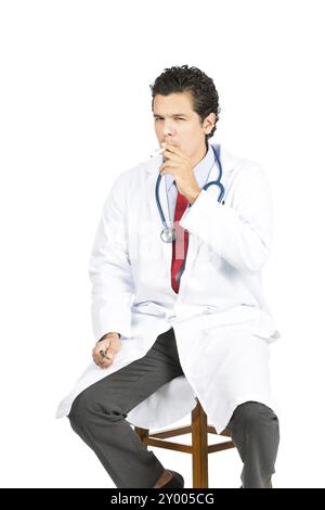 Un bel medico latino, che indossa il bianco, che dimostra una pratica malsana fumando e inahling profondamente il fumo di sigaretta, guardando la macchina fotografica. V Foto Stock