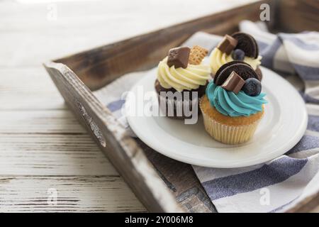 Gustosi muffin multicolore e cupcake sul piatto bianco su un vassoio di legno con asciugamano. Decorate con caramelle, biscotti e croccanti colorati Foto Stock