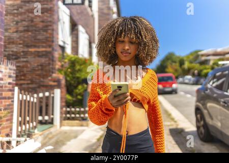 Vista frontale di una bella donna latina con capelli ondulati e vestiti informali colorati usando il telefono che cammina lungo un quartiere residenziale Foto Stock