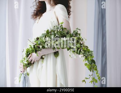 Crop donna a piedi nudi in elegante abito da sposa bianco con grande bouquet di piccoli fiori bianchi e verde fogliame in piedi tra trasparente colore pastello c Foto Stock