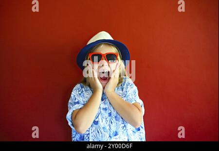 Piuttosto emothional bambino indossare un cappello e occhiali da sole su uno sfondo di colore rosso. Copia spazio, luce diurna Foto Stock