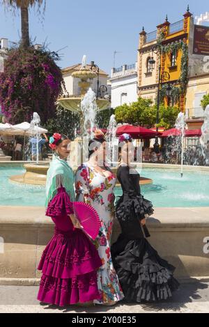 Tre donne in abiti flamenco colorati, con fiori nei capelli, stanno di fronte a una fontana in una piazza della città sotto la luce del sole, Feria de la Manza Foto Stock