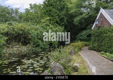 Uno stretto sentiero lungo una sponda del fiume con ninfee e una casa in mattoni sullo sfondo, aalten 2 Foto Stock