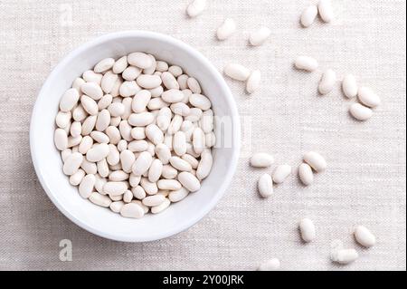 Fagioli bianchi in una ciotola bianca su tessuto di lino. Semi piccoli essiccati, una varietà di fagioli comuni Phaseolus vulgaris. Foto Stock