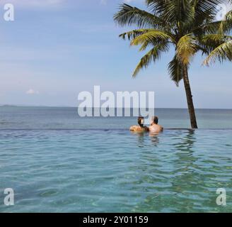 Coppia giovane rilassante nella piscina a sfioro sotto le palme di cocco nella parte anteriore del paesaggio tropicale Foto Stock