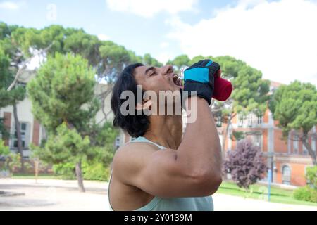 Un uomo sta bevendo acqua da una bottiglia mentre indossa i guanti. La scena è ambientata in un parco con alberi e una panchina. Quell'uomo si sta prendendo una pausa dal suo lavoro Foto Stock