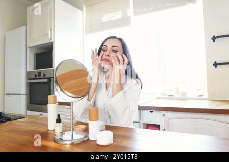 Carino donna sorridente l'applicazione di crema per il viso Indossare tunica bianca al mattino presso la cucina Foto Stock