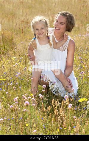 La bella giovane madre e la sua dolce bambina vestite con abiti estivi bianchi, sedute su un prato con fiori selvatici in una calda giornata estiva Foto Stock