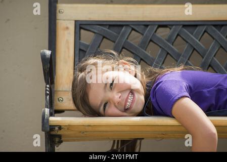 Bambina sdraiata sullo stomaco su una panchina esterna con un grande sorriso sul viso Foto Stock