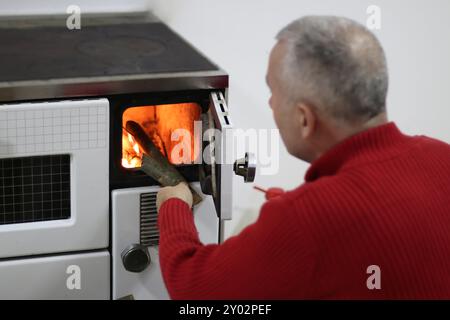 Povero uomo maturo che mette la legna nella stufa all'interno a casa, concetto di povertà. Foto Stock