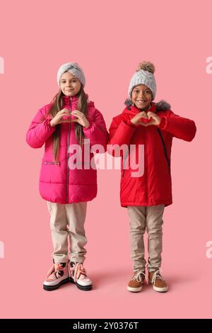 Bambini piccoli e felici con abiti invernali che mostrano gesti del cuore su sfondo rosa Foto Stock