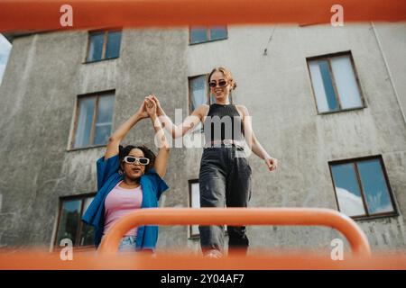 Giovane donna che tiene per mano un'amica donna che si mette in equilibrio su una barra di metallo nel parco giochi contro l'edificio Foto Stock