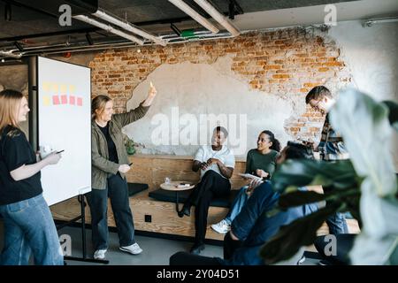 Programmatore tecnologico donna che mostra una nota adesiva ai colleghi felici durante una riunione d'affari in ufficio Foto Stock