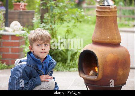 Simpatico ragazzo biondo seduto a terra con la sua borsa a mano sulla schiena guardando la macchina fotografica di fronte a un tradizionale bruciatore a legna da esterno con un burni Foto Stock