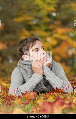 Giovane donna attraente sdraiata in foglie d'autunno colorate, con la testa appoggiata sulle braccia. Espressione facciale premurosa. Una donna attraente stesa sullo stomaco Foto Stock