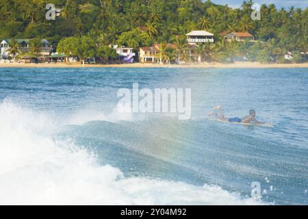 Un piccolo arcobaleno si forma come surfista non identificabile dello Sri Lanka e' visto pagaiare su un'onda al largo della spiaggia di Unawatuna, un luogo di surf notevole in Sri Lanka Foto Stock