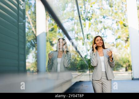 Una donna professionista cammina con facilità, sorridendo mentre si concentra sul telefono, mescolando lo stile di vita moderno con la tranquillità della natura sullo sfondo Foto Stock