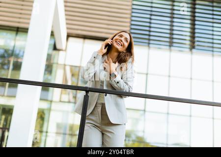 Una donna d'affari gioiosa festeggia un momento all'aperto, partecipando a una allegra conversazione telefonica mentre si appoggia a una ringhiera accanto a un elegante buildi Foto Stock