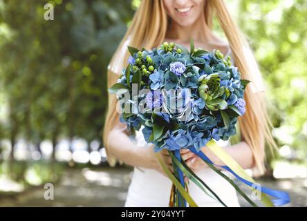 Crop sorridente giovane bionda dai capelli lunghi femmina che tiene bella bouquet elegante di fiori verde e blu con nastri mentre si trova in parco con garee Foto Stock