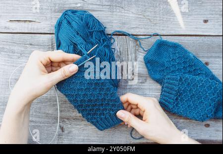 Donna che tiene in mano il lavoro a maglia blu su fondo di legno grigio. Ragazza a maglia in giardino. Uncinetto a maglia blu in mani da ragazza su un tavolo di legno. Foto Stock