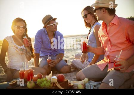 Gruppo di giovani amici allegri e alla moda che bevono succo di frutta mentre si picnic e godersi il tempo insieme sulla spiaggia di sabbia nella soleggiata giornata estiva Foto Stock