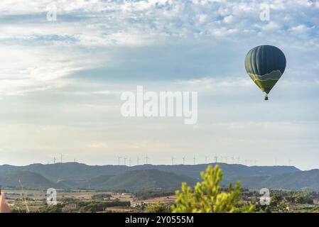 Mongolfiera che sorvola un paesaggio panoramico con turbine eoliche sullo sfondo, sotto un cielo parzialmente nuvoloso Foto Stock