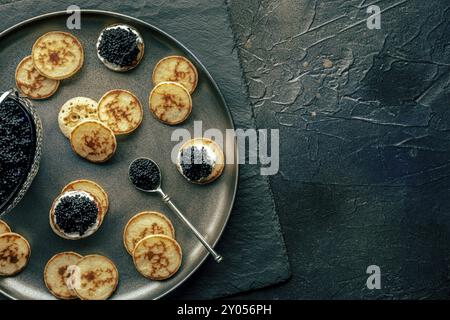 Blinis con caviale nero e formaggio cremoso, piatto sopra la testa su sfondo nero, mini pancake, un elegante antipasto, con spazio per copiare Foto Stock