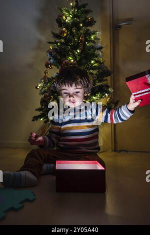 Un bambino è seduto davanti a un albero di Natale e ha in mano una scatola rossa. Sta aprendo la scatola e la scena è ambientata in una stanza poco illuminata. La scena è f Foto Stock