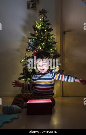 Un ragazzino è seduto di fronte ad un albero di Natale, aprendo una scatola rossa. La scena è gioiosa e festosa, mentre il ragazzo è circondato dalle festività natalizie Foto Stock