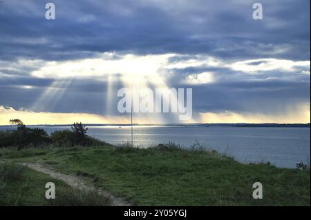 Sulla costa di Hundested. I raggi del sole si rompono attraverso il cielo drammatico attraverso le nuvole. Prato con sentiero in primo piano. Paesaggio girato in Danimarca Foto Stock