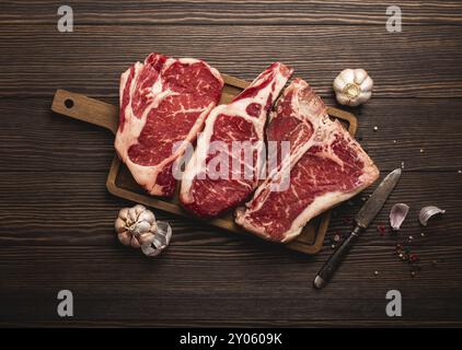 Insieme di tre tipi diversi di bistecche di carne cruda: Ribeye, T-bone, cowboy su tavola tagliata con coltello e condimenti, fondo in legno. Assortimento di bistecche invecchiate Foto Stock