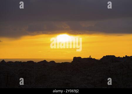 Silhouette of two people standing on distant ridge watching sun breaking thru clouds making a colorful orange sky at sunset in Gobi Desert in rural Mo Stock Photo