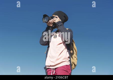 Ritratto di una ragazza hippy elegante con un cappello e uno zaino che la fotografa con una fotocamera reflex digitale all'aperto contro un cielo blu in una zona soleggiata Foto Stock