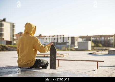 Primo piano di un giovane hipster vestito con una felpa e jeans seduti tristi in uno skate Park e con in mano uno skateboard Foto Stock