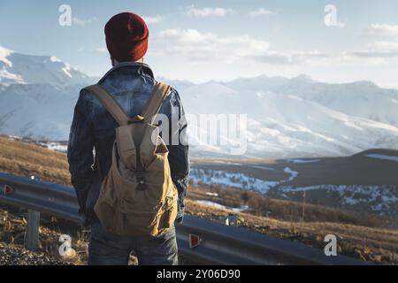 Uomo hipster turistico barbuto con occhiali da sole e zaino, stenditi su un dosso lungo la strada e guarda il tramonto sullo sfondo di una cima innevata Foto Stock