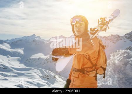 Ritratto di uno sciatore in una tuta arancione con uno zaino sulla schiena e gli sci sulle spalle in un casco si erge su una roccia contro il cielo blu. Copia Foto Stock