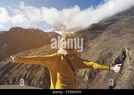 Una giovane donna viaggiatrice con un grande cappello di pelliccia e un maglione in maglia gialla è in piedi con le braccia allungate sulle montagne sullo sfondo di t Foto Stock