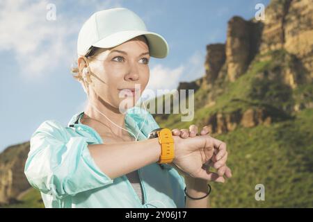 Ritratto di una giovane ragazza del fitness sorridente con un berretto e le cuffie che controllano il suo orologio intelligente mentre si siede all'aperto su uno sfondo di rocce. Guarda Foto Stock